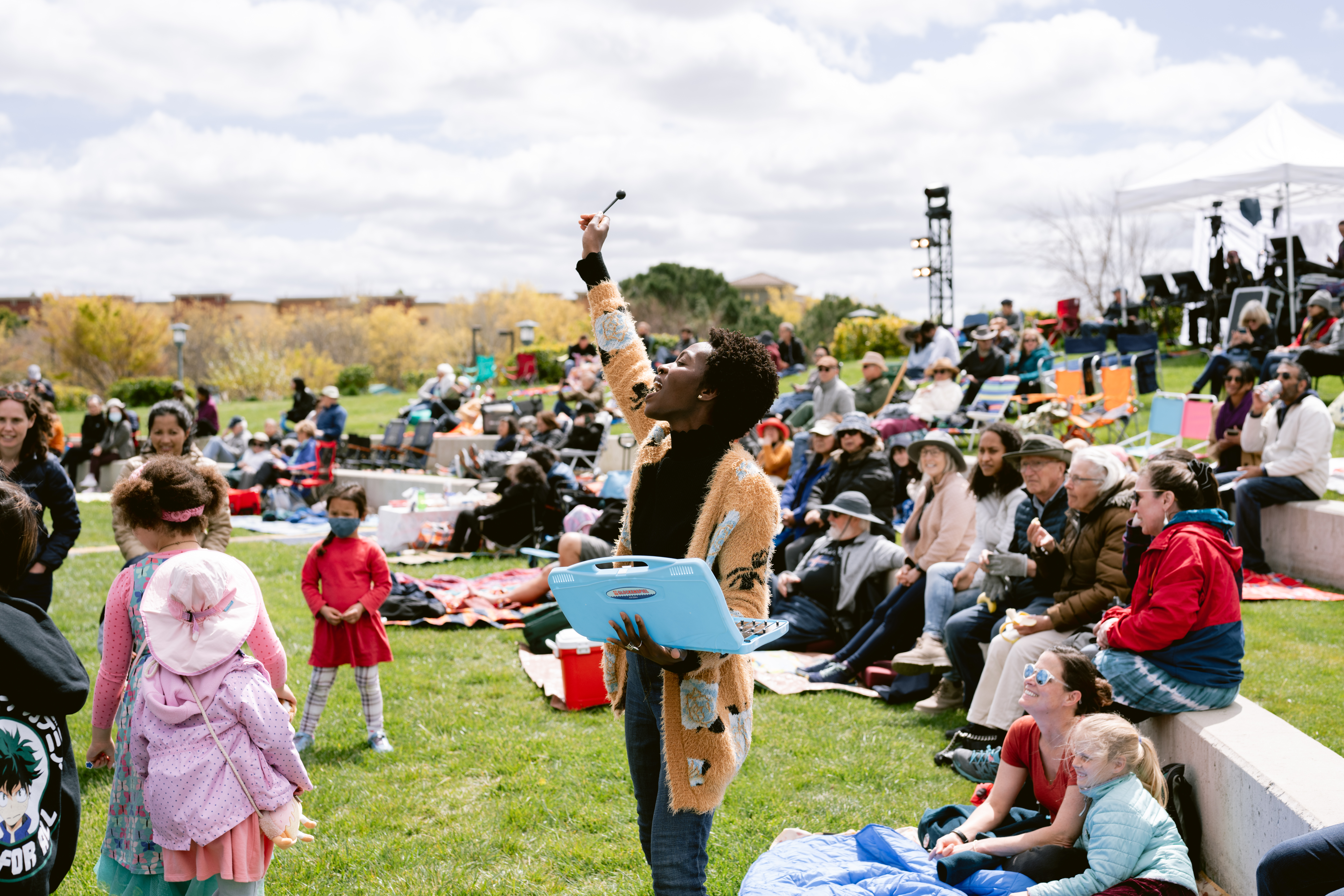 A photo from an outdoor event with people sitting on the lawn. A woman is standing, holding a laptop, raising her hand in the air.